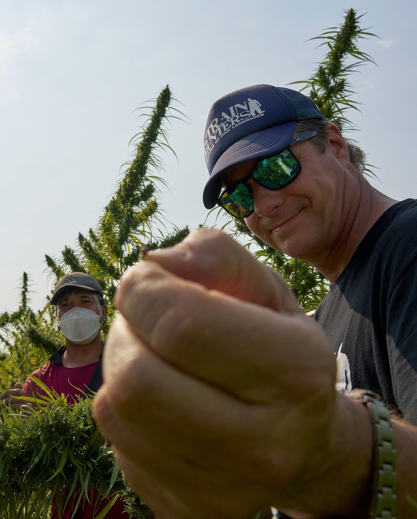 A single seed held between finger and thumb in the foreground with expedition workers and plants behind